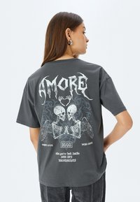 Camiseta de gran tamaño en gris oscuro con gráficos de calaveras en blanco, diseño de alas intrincado y texto. Fabricada en una mezcla de algodón con un corte relajado.