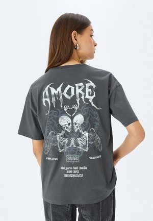T-shirt oversize grigio scuro con grafiche di teschi bianche, design di ali intricati e testo. Realizzato in misto cotone con una vestibilità comoda.