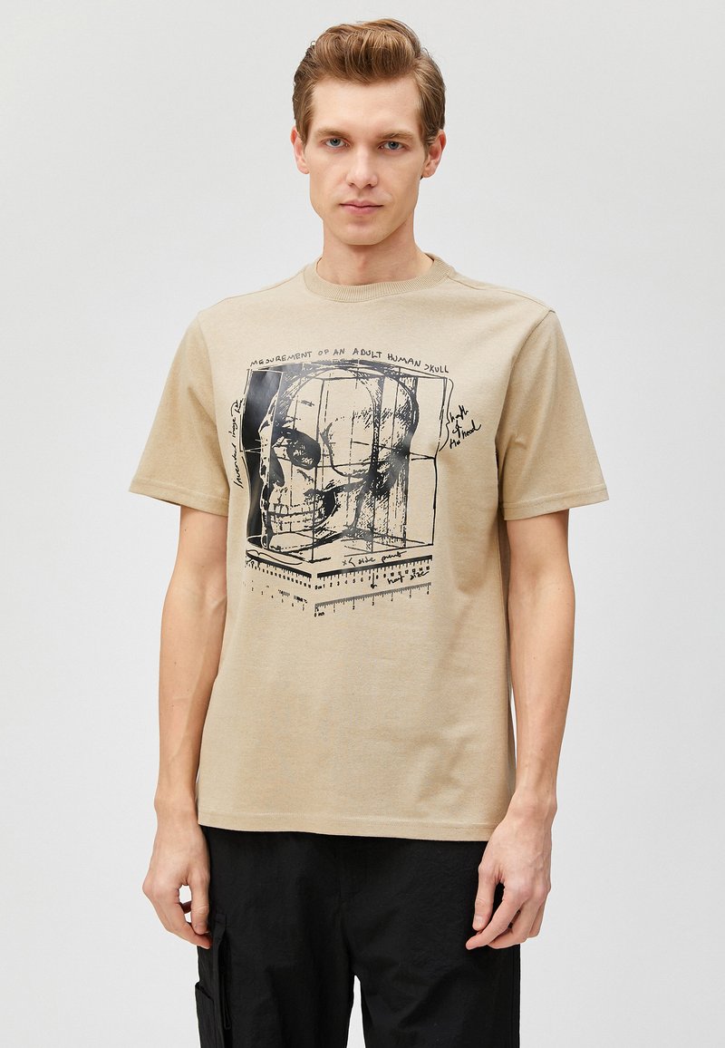 Camisa de algodón beige con un estampado gráfico negro de una calavera con líneas de medida y texto escrito a mano en la parte frontal. Mangas cortas, cuello redondo.