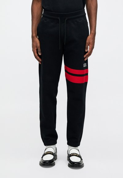 Svarta sweatpants med röda ränder på sidan, dragsko i midjan och en liten logo. Tillverkade av ett mjukt material med en lätt strukturerad yta.