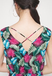 Kvinna med mörkt hår i hästsvans som bär stora hoops örhängen och en klänning med tropiskt mönster och tunna korsade band bak.