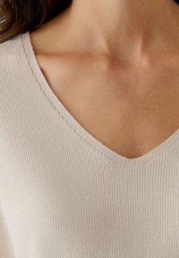 Helle beige, gerippte Strickjacke mit V-Ausschnitt, die eine weiche Textur und minimale Nähte am Kragen aufweist.