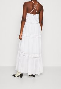 Bílá maxi šaty s rozšířeným lemem, akcenty z krajky a nastavitelnými křížovými ramínky. Hladká textura a lehká tkanina. Černé podpatky.