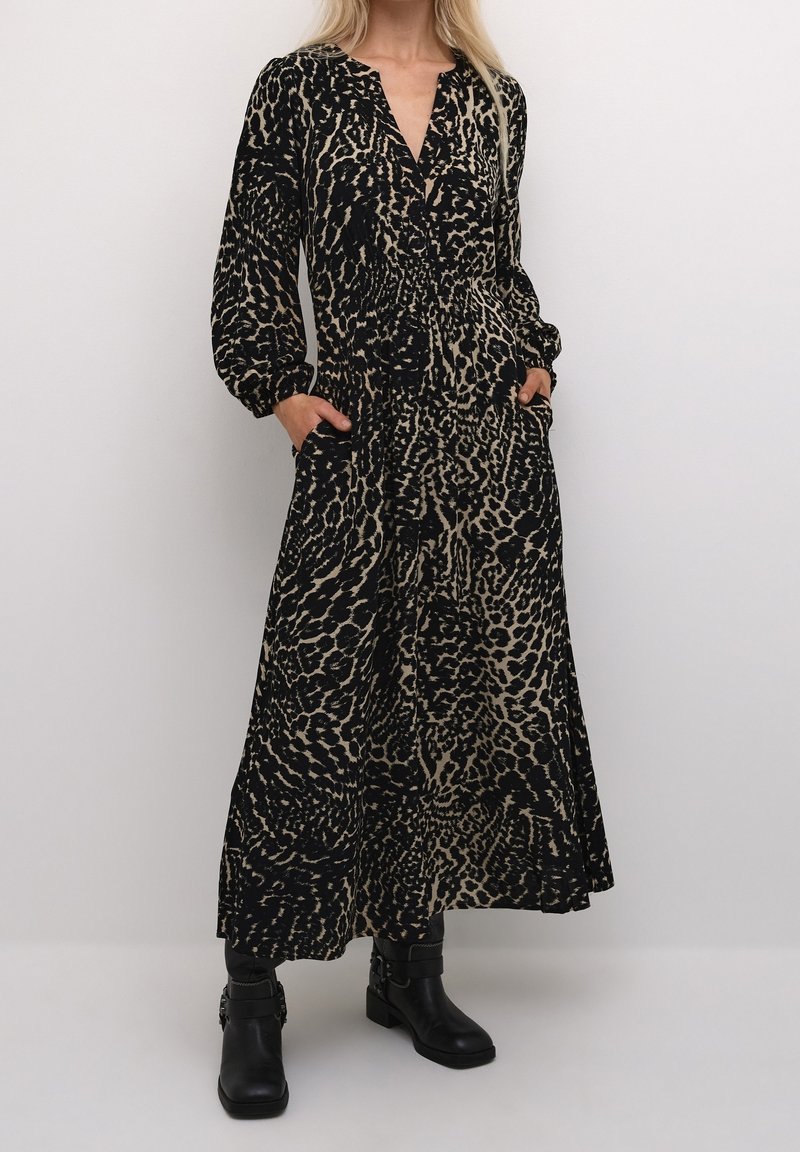 Femme portant une longue robe à imprimé animal noir et beige avec des manches longues et des bottines noires, debout les mains dans les poches.