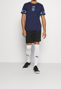 Man i marinblå Puma Manchester City fotbollsjacka, svarta shorts, vita Puma knästrumpor och svarta sneakers, står.