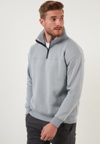 Sudadera de color gris claro con cuello de embudo con cremallera, puños acanalados y una textura suave, que presenta una costura horizontal en el pecho.