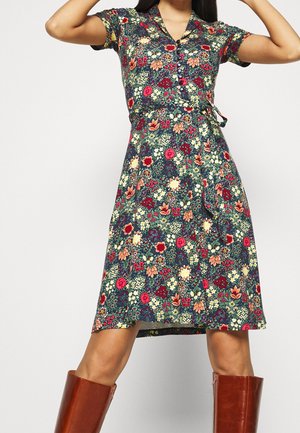 Kobieta ubrana w sukienkę w kwiaty do kolan z krótkimi rękawami, z guzikami i paskiem w talii, zestawioną z brązowymi kozakami za kolano.