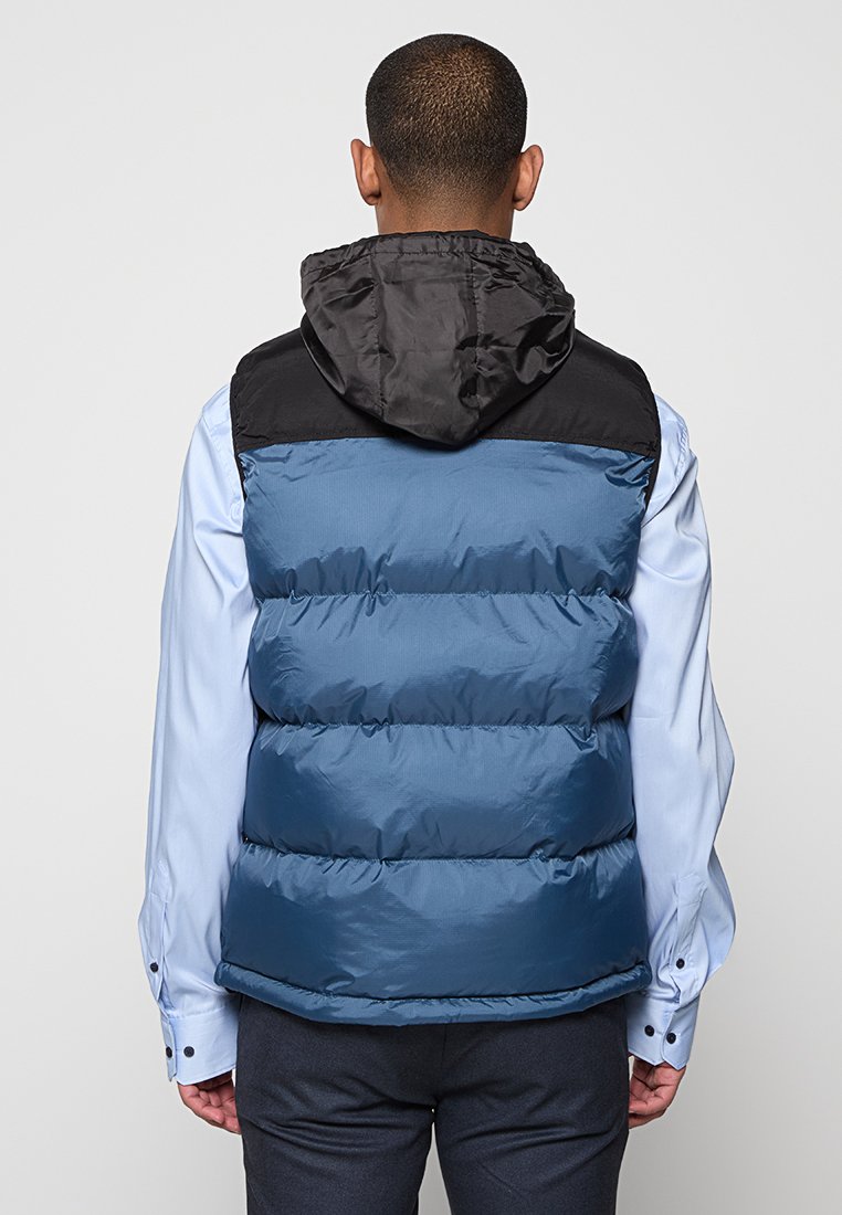 Doudoune sans manches avec une capuche noire, présentant un corps texturé bleu et un motif matelassé horizontal. Portée sur une chemise bleu clair, avec un pantalon foncé.