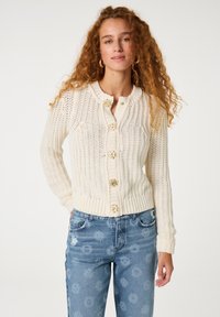 Gehaakte crème cardigan met een ronde halslijn, geribde manchetten en zes decoratieve bloemenknopen. Gedragen over lichtblauwe, gedessineerde jeans.