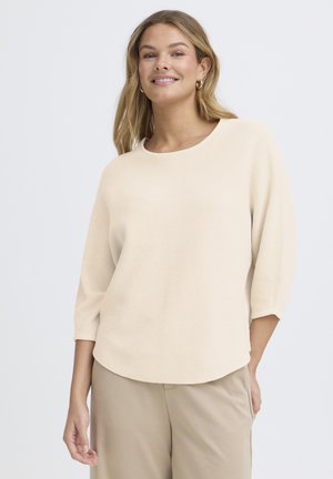 Donna sorridente con lunghi capelli biondi che indossa un maglione crema a coste con maniche a tre quarti e pantaloni beige, in piedi contro uno sfondo bianco.