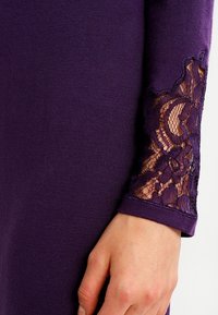 Abito viola con maniche lunghe e dettagli in pizzo sui polsini. Il tessuto sembra liscio, mentre il pizzo presenta un intricato motivo floreale.