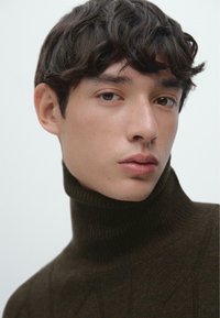 Maglione dolcevita marrone scuro a coste con motivo testurizzato. Primo piano di un modello maschile con capelli scuri spettinati e carnagione naturale.