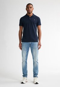 Polo shirt blu navy con un piccolo logo, abbinato a jeans azzurri. L'outfit include sneakers bianche, con una tomaia dalle texture e dettagli neri.