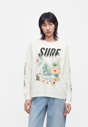Ung person med sort hår iført en hvid sweatshirt med "SURF" tekst, blomster- og bølgemønster samt lyseblå jeans, stående mod en hvid baggrund.