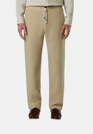 Pantaloni in lino beige con vita regolabile con coulisse, indossati con camicia beige chiaro e mocassini in pelle marrone scuro su sfondo bianco.
