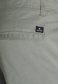 Gros plan sur la poche arrière d'un pantalon gris clair avec une étiquette de marque noire Jack & Jones au-dessus de la poche fendue horizontale.