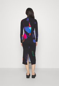 Femme portant une robe midi noire à manches longues avec un motif abstrait coloré et une fente à l'arrière, associée à des sandales compensées noires.
