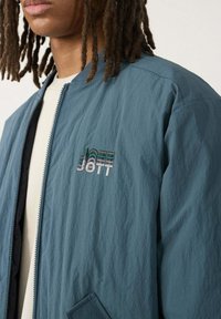 Giacca bomber verde acqua con finitura testurizzata, dotata di zip frontale e tasca laterale. Il logo "JOTT" è ricamato con fili multicolori.
