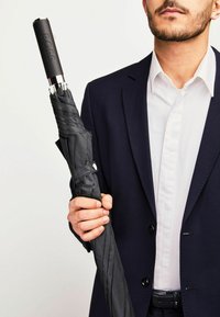Parapluie noir à motifs avec une poignée texturée, arborant le logo "BOSS", tenu par une personne en costume sombre et chemise blanche.