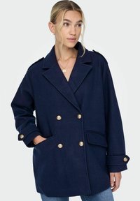 Manteau navy à double boutonnage en mélange de laine, avec des boutons dorés, de grandes poches et un col structuré. Coupe décontractée avec des manches longues.