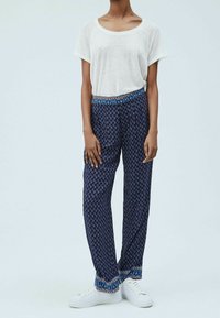 Marineblau gemusterte Hose mit einem blauen Taillenbund und einem weißen T-Shirt mit Grafik, kombiniert mit weißen Turnschuhen vor einem hellen Hintergrund.