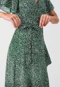 Vestido verde con puntos blancos, escote en pico, mangas cortas con vuelo, cintura atada y falda fluida hecha de una tela suave y texturizada.