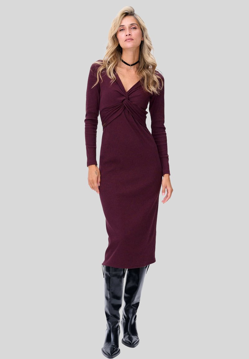 Bordowa dzianinowa sukienka z skręconym przodem, długimi rękawami i dopasowanym krojem, zestawiona z czarnymi błyszczącymi kozakami do kolan.