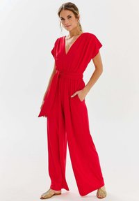 Czerwony kombinezon z głębokim dekoltem w kształcie litery V, krótkimi rękawami i wiązaniem w talii. Luźny, szeroki krój nogawek z bocznymi kieszeniami. Gładki, lekki materiał.