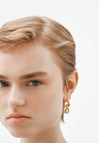 Pendientes de metal dorado con un diseño de bucle retorcido y acabado pulido, colgando de la lóbulos de la oreja. El fondo es neutro y suave.