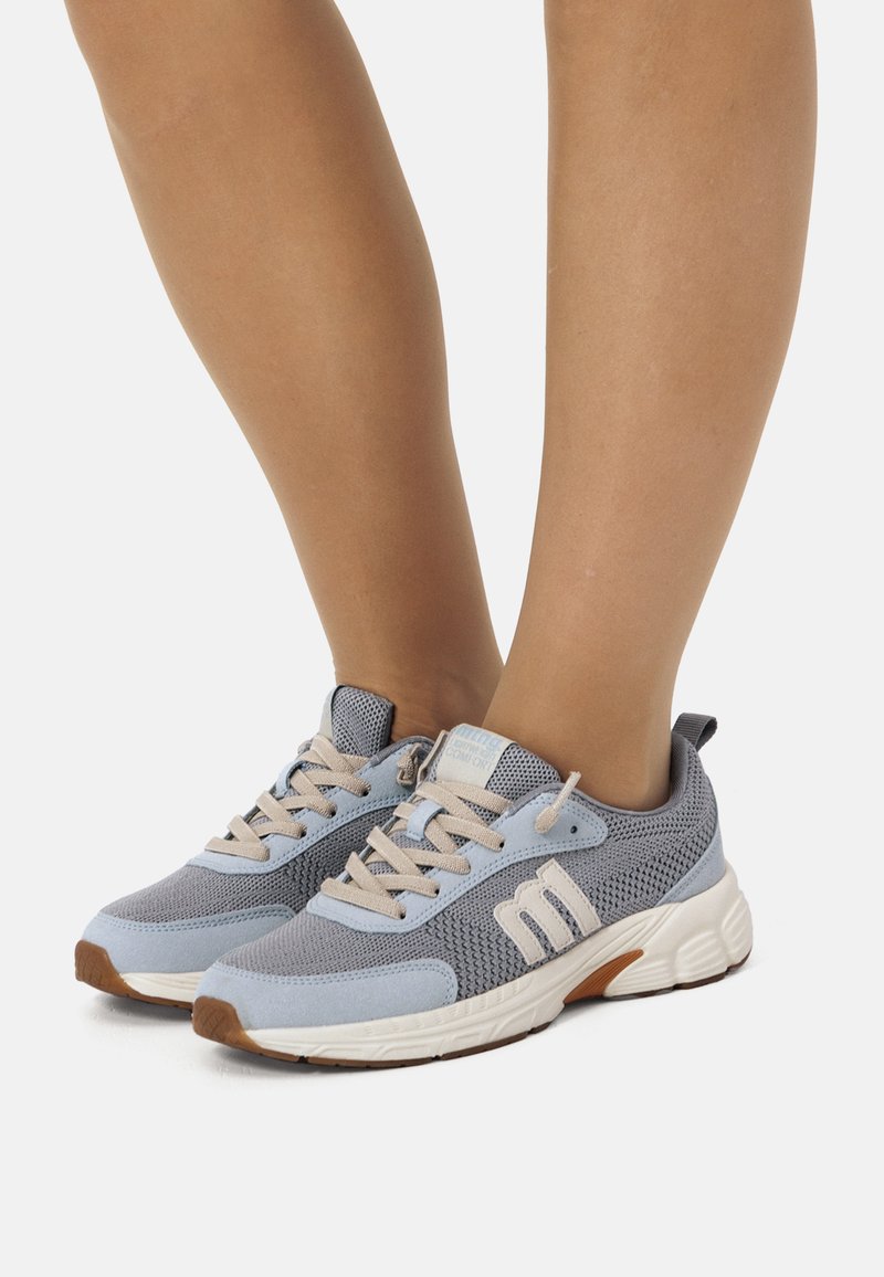 Piernas vestidas con zapatillas de malla en azul claro y gris, con cordones beige y suelas blancas gruesas, de pie sobre una superficie blanca.