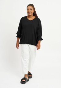 Top nero a V con maniche lunghe e puffate, con polsini arricciati; abbinato a pantaloni bianchi a vestibilità ampia e sandali neri con nodo.