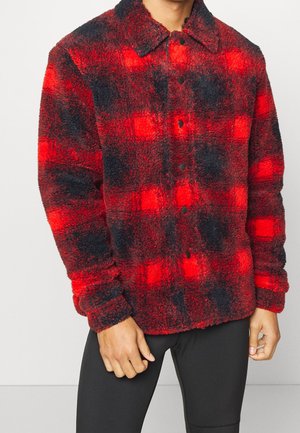 Mężczyzna noszący czerwoną i czarną polarową kurtkę w kratę z kołnierzykiem i czarnymi guzikami, w połączeniu z czarnymi spodniami.