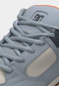 Nahaufnahme eines hellgrauen Sneakers mit strukturierten Schnürsenkeln, perforierter Seitenpartie und DC-Markenlogo auf der Zunge mit orangefarbener Umrandung.