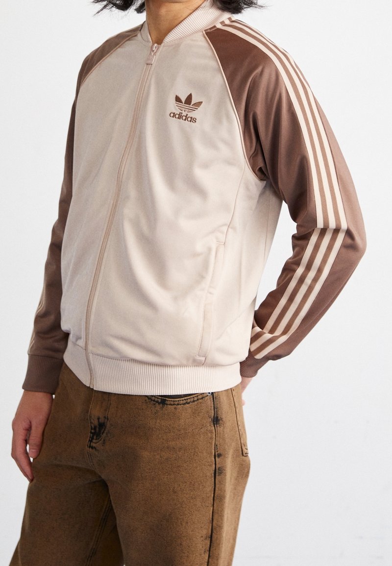 Person wearing beige and brown Adidas track jacket with three stripes on sleeves and brown jeans, hands relaxed by sides.