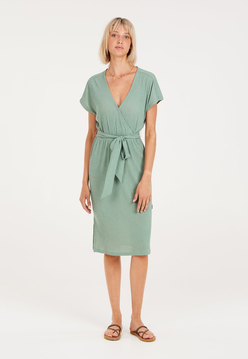 Vestido verde con manga corta, cinturón de lazo, tejido texturizado y faldón hasta la rodilla. Llevado con sandalias planas, con una sutil abertura lateral.