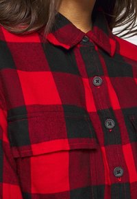 Camicia in flanella a quadri rossi e neri con chiusura a bottoni, colletto e due tasche frontali con bottoni neri. Tessuto morbido.