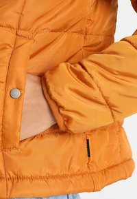 Vadderad orange jacka med en blank yta, som har en snug passform runt handleden, en sidoficka och detaljer med silverknapp.