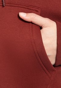 Felpa color marsala con una texture liscia; presenta una tasca frontale con una mano inserita, che mette in risalto i dettagli dell'imbastitura e dei bordi.