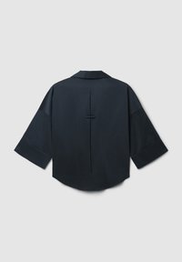 Camicia oversized blu scuro con maniche ampie, una piega centrale sul retro e un piccolo dettaglio a loop orizzontale in tessuto su uno sfondo chiaro.