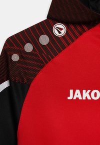 Rotes Sporttrikot mit schwarzen Akzenten, strukturiertem Streifenmuster an der Schulter, weißem Logo "JAKO" und drei runden Designelementen.
