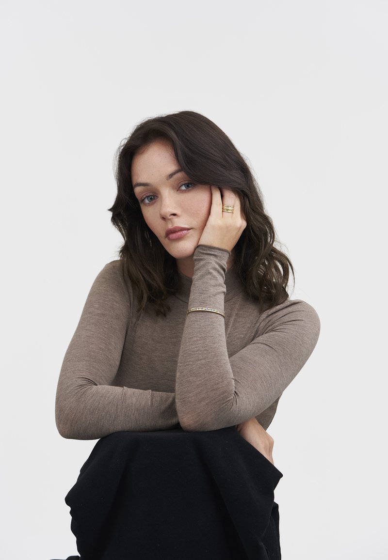 Woman with dark wavy hair wearing a brown long-sleeve top and gold bracelet and rings, resting her face on her left hand against a white background.