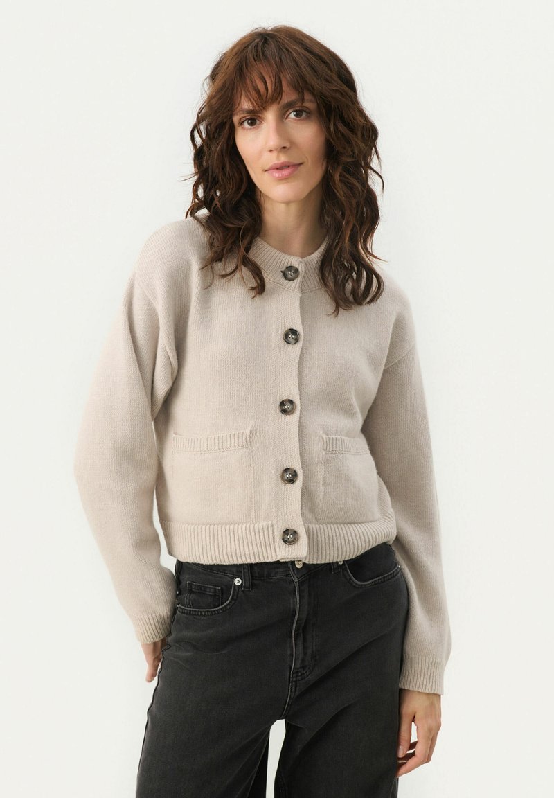 Cardigan en maille beige avec un col rond, poches avant et six boutons foncés ; ourlet et poignets côtelés ; texture lisse.