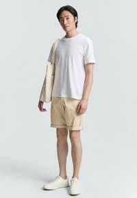 Beige Shorts, weißes T-Shirt und weiße Sneaker. Die Person hält eine beige Tragetasche. Minimalistisches Design mit einem lässigen Look.