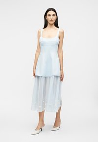 Femeie cu păr lung și închis la culoare, purtând o rochie fără mâneci de culoare albastru deschis, cu fustă transparentă brodată cu modele florale și pantofi albi cu toc.