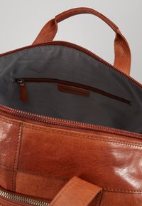 Sac en cuir brun avec une ouverture zippée, présentant une doublure à carreaux gris et noirs et une poche zippée intérieure pour l'organisation.