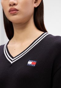 Μαύρο πουλόβερ με V λαιμόκοψη και λευκές ρίγες στις μανσέτες. Διαθέτει ένα πολύχρωμο λογότυπο Tommy Jeans στο μπροστινό μέρος.