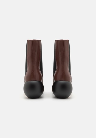 Des bottes en cuir marron à la cheville avec des côtés en tissu extensible noir, un talon en caoutchouc noir et des détails de couture verticaux à l'arrière.