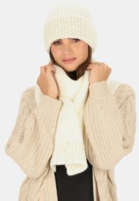 Biały prążkowany zestaw czapki beanie i szalika, charakteryzujący się miękką teksturą i przytulnym designem, noszony na tle swetra w neutralnych odcieniach.