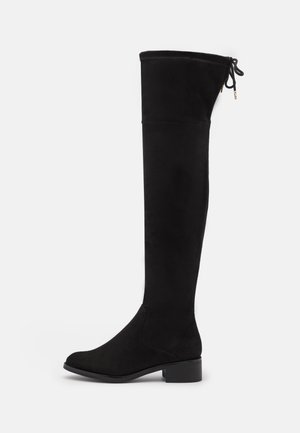 Botte plate noire au-dessus du genou avec un petit talon bloc et un détail nœud noué sur le haut du dos, présentée sur fond blanc.