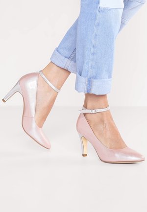 Stopy w jasnoniebieskich, podwiniętych dżinsach, ubrane w połyskujące różowe szpilki z paskami wokół kostki na jasnym tle.